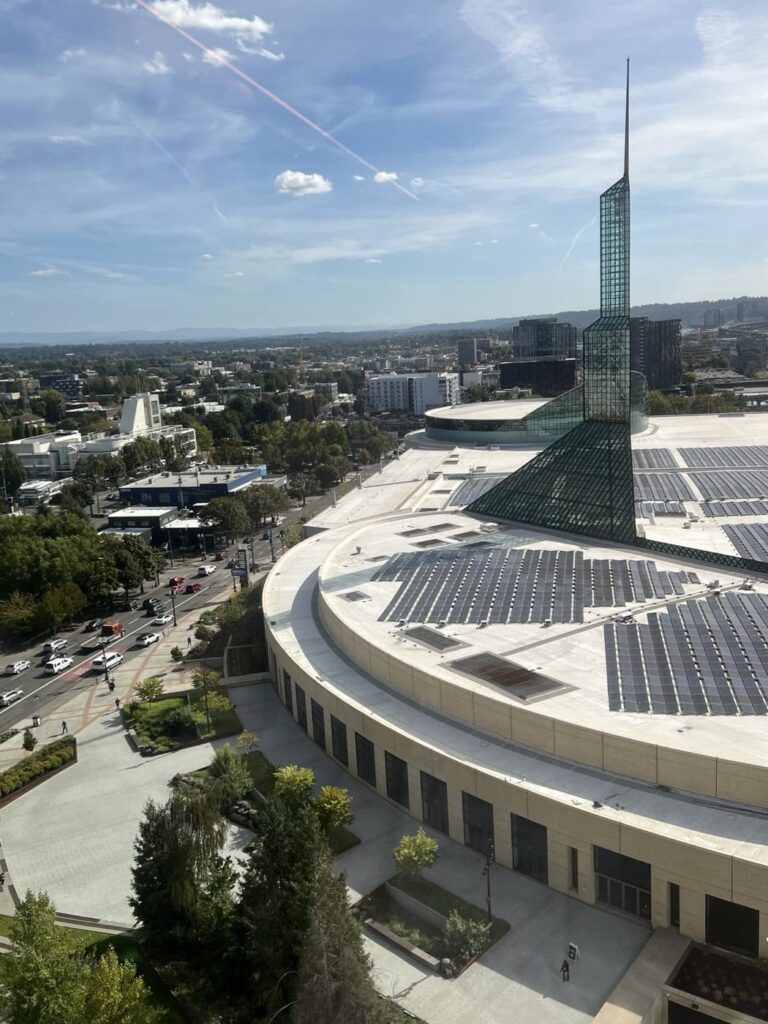 Aerial view of the Oregon Convention Center. The structure has a distinctive spire and solar panels on its curved roof. Surrounding the structure are roads, trees, and other buildings under a partly cloudy sky.