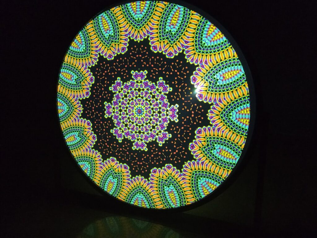 A circular kaleidoscope pattern with vibrant colors, including green, yellow, orange, and purple, forming intricate symmetrical designs against a black background, creating a visually mesmerizing effect.