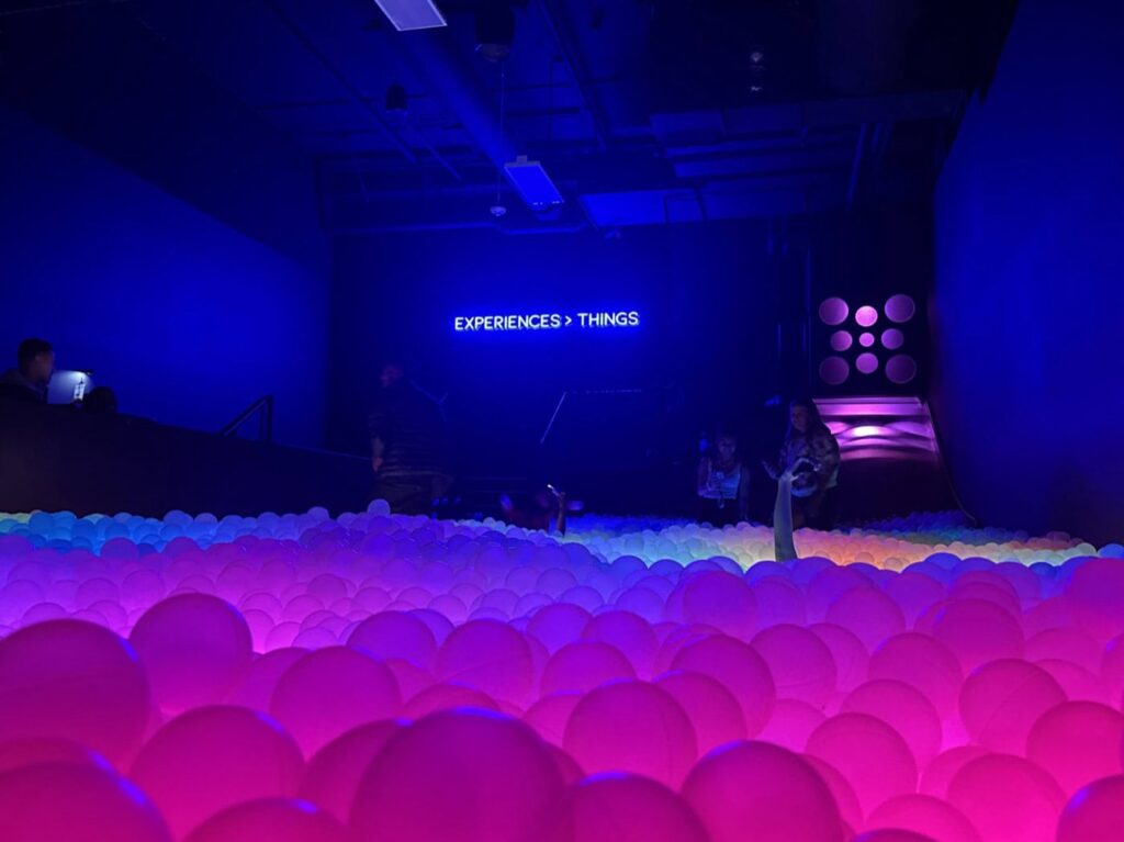 A dimly lit room with people exploring a wide ball pit filled with glowing pink and purple balls. A neon sign on the back wall reads EXPERIENCES > THINGS. The atmosphere is futuristic and immersive.