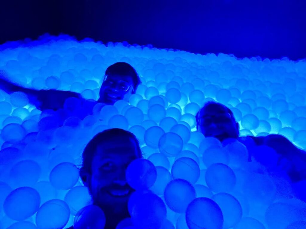 Vinícius, Max and Scott immersed in a glowing blue ball pit, smiling and enjoying the experience. The lighting gives the scene a surreal and playful atmosphere.
