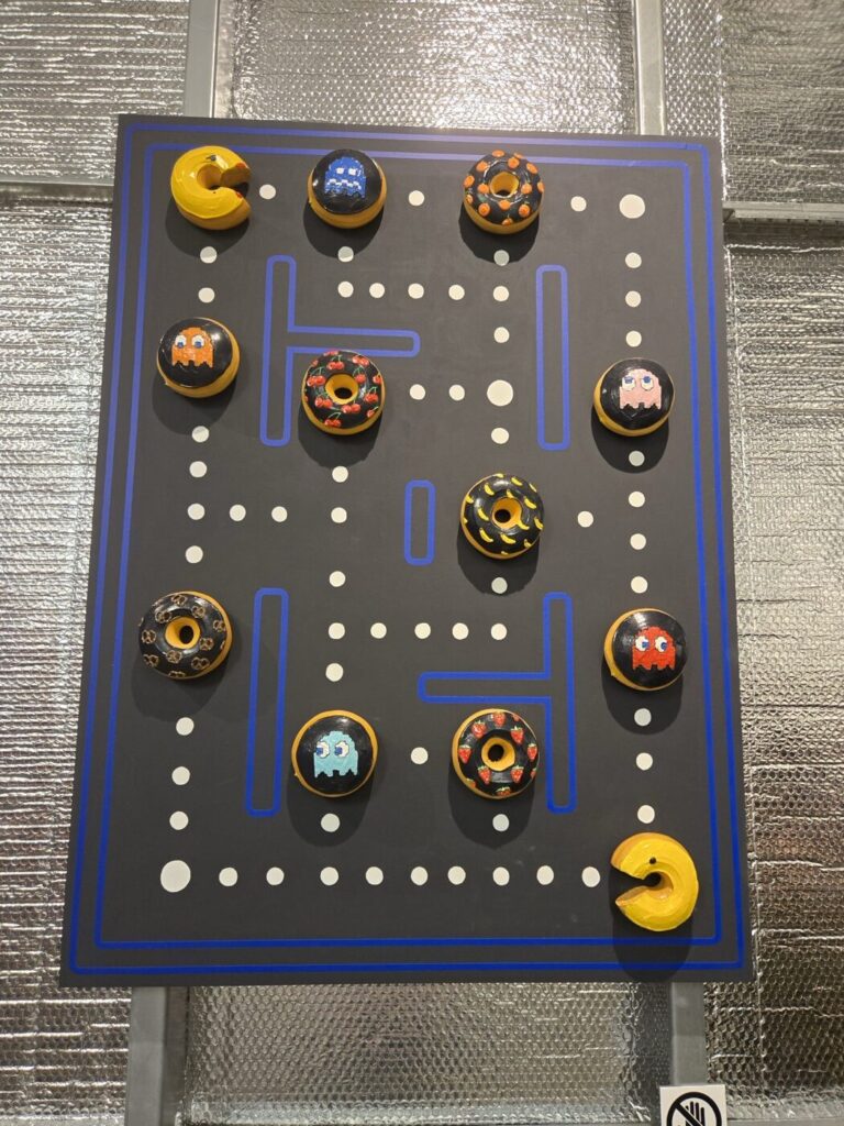 A wall display featuring a Pac-Man-themed design, where donuts are creatively placed to resemble game elements such as ghosts and Pac-Man himself. The background mimics the classic game's maze with paths and dots.