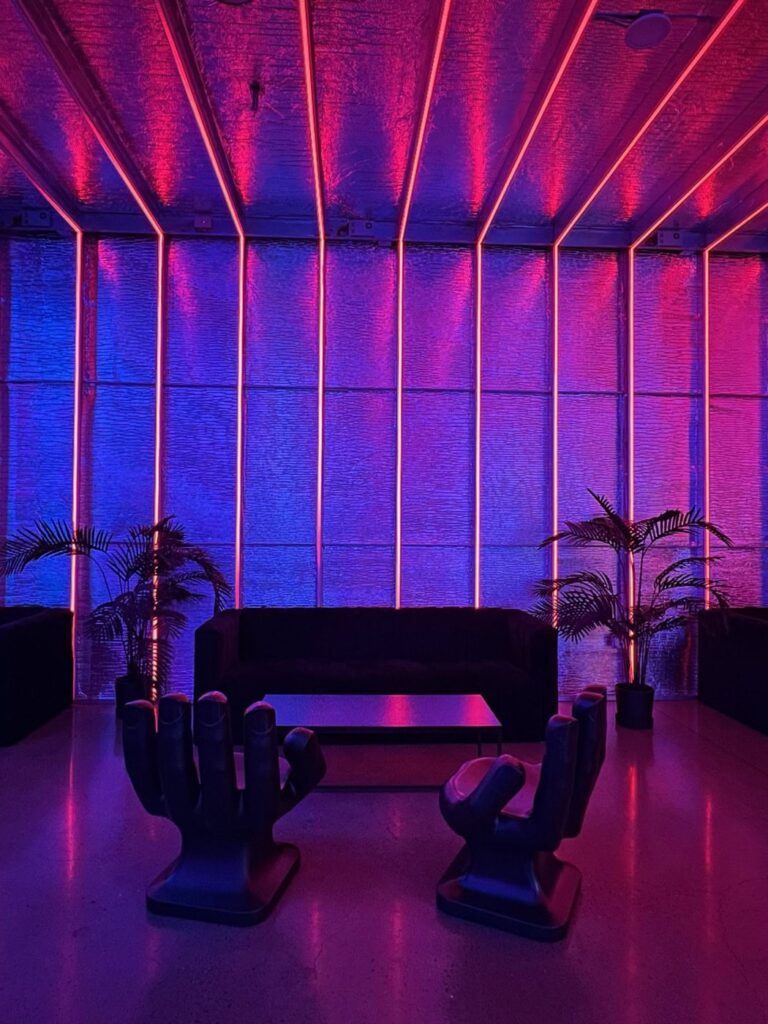 A modern lounge with vibrant pink and blue lighting. It features a sleek black sofa, a coffee table, two unique hand-shaped chairs, and potted plants, creating an atmospheric and stylish setting.