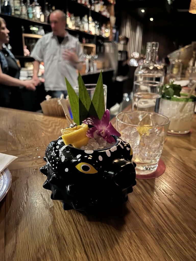 A creative cocktail presentation in a black, spiky pufferfish mug at Eem Thai, garnished with green leaves, purple flowers, and lemon slices. The drink rests on a wooden bar counter, with a blurred background of a bar and two people.