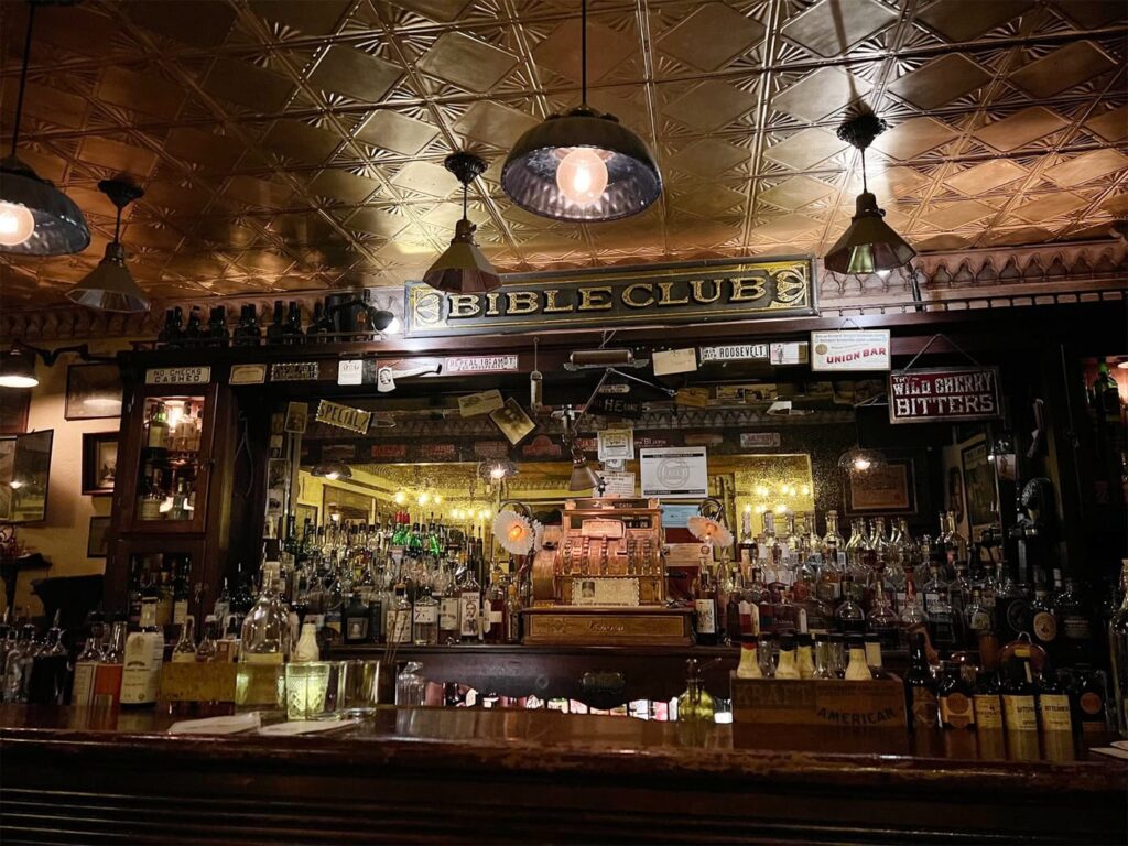 Bible Club's cozy prohibition-era bar interior with a vintage vibe featuring a counter lined with various bottles of spirits. A sign that reads "Bible Club" is prominently displayed above the bar, surrounded by warm lighting, decorative hanging lamps and a gold plated ceiling.