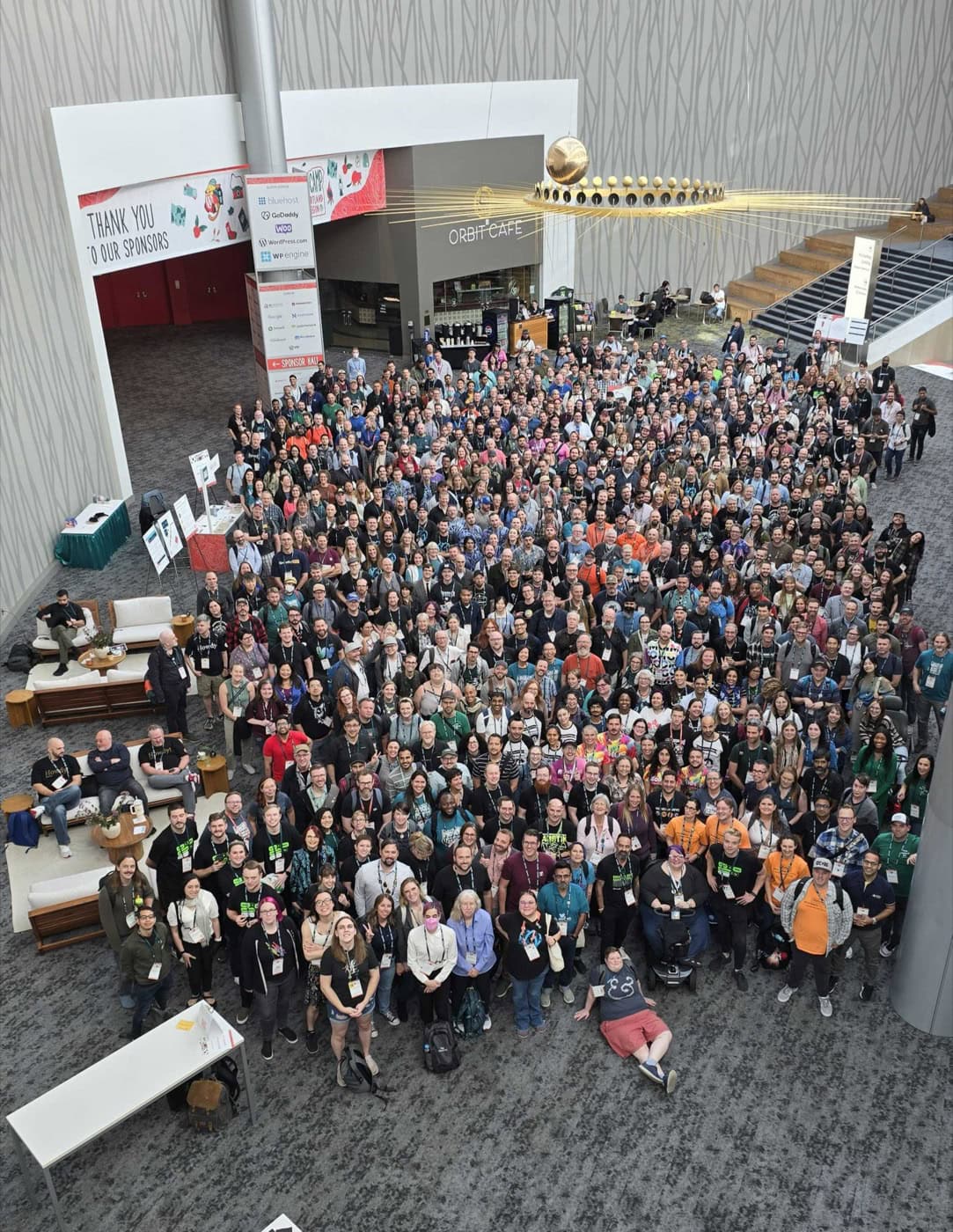 A large number of conference attendees gather in the Oregon Convention Center's spacious lobby, posing for a group photo. The crowd is diverse, with various ages and styles of dress. Some are seated on couches while most are standing.