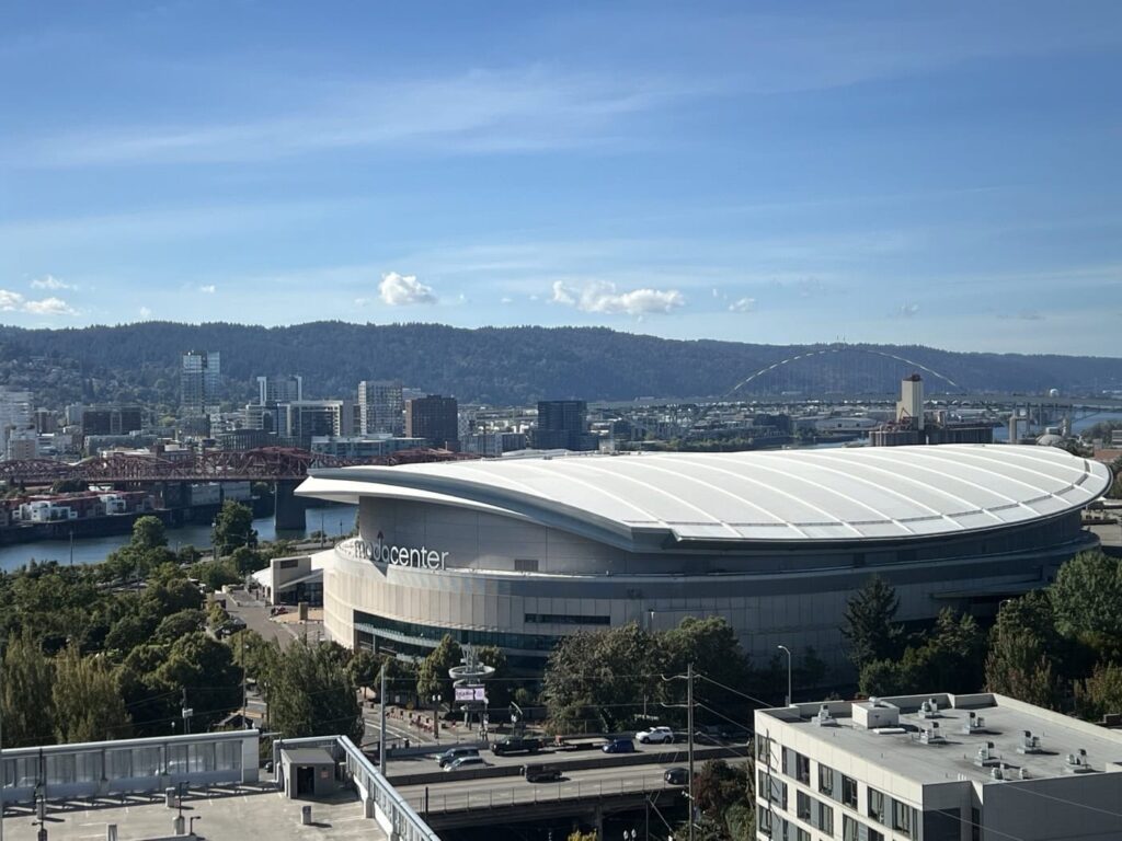 Aerial view of a cityscape featuring the Moda Center, a large modern arena with a white curved roof. The arena is surrounded by trees, roads, and buildings, with a river and bridge in the background under a clear blue sky.