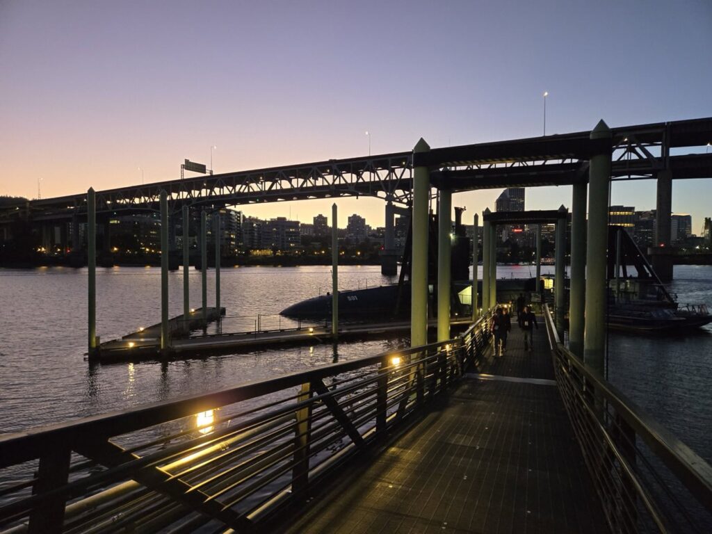 A dock at twilight with a view of a bridge spanning a calm river. Soft lighting creates reflections on the water, and city buildings are silhouetted against the sky. A few people are walking towards a docked submarine.