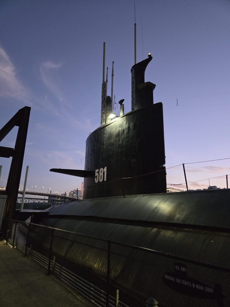The USS Blueback submarine is docked with a twilight sky behind. The vessel's conning tower and periscopes are visible against a darkening sky, with faint lights in the background. A fence and signage are in the foreground.