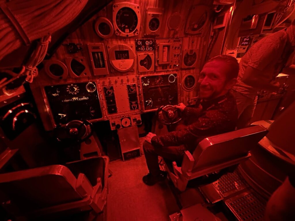 Max sitting in the dimly lit cockpit holding the steering wheel, surrounded by various control panels and gauges with red lighting.