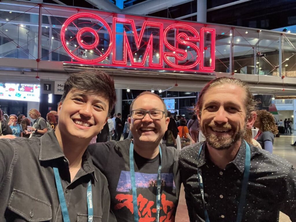 Vinícius, Scott and Max smile in front of a large neon red "OMSI" sign at the WordCamp US afterparty. Other Wordcamp attendees are socializing in the background.