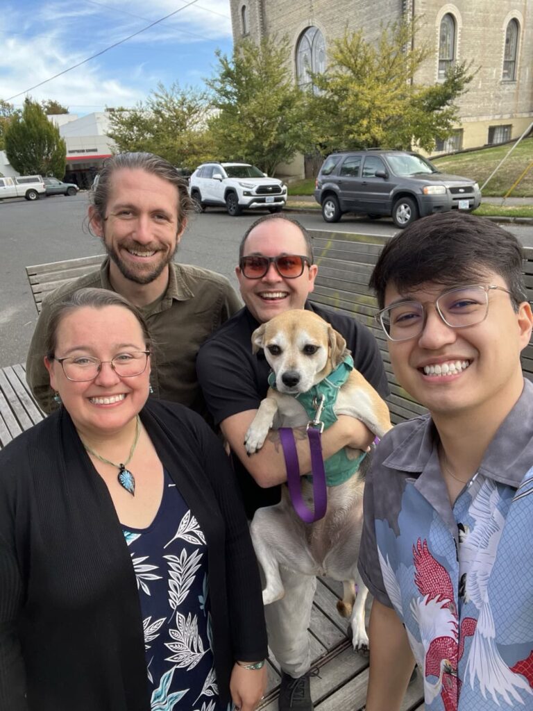 Max, Scott, Lindsey and Vinícius smiling at the camera while standing outside of Pine State Biscuits. Scott is holding Mabel, a tan and white dog with a green harness and purple leash. There are buildings, trees and vehicles visible in the background.
