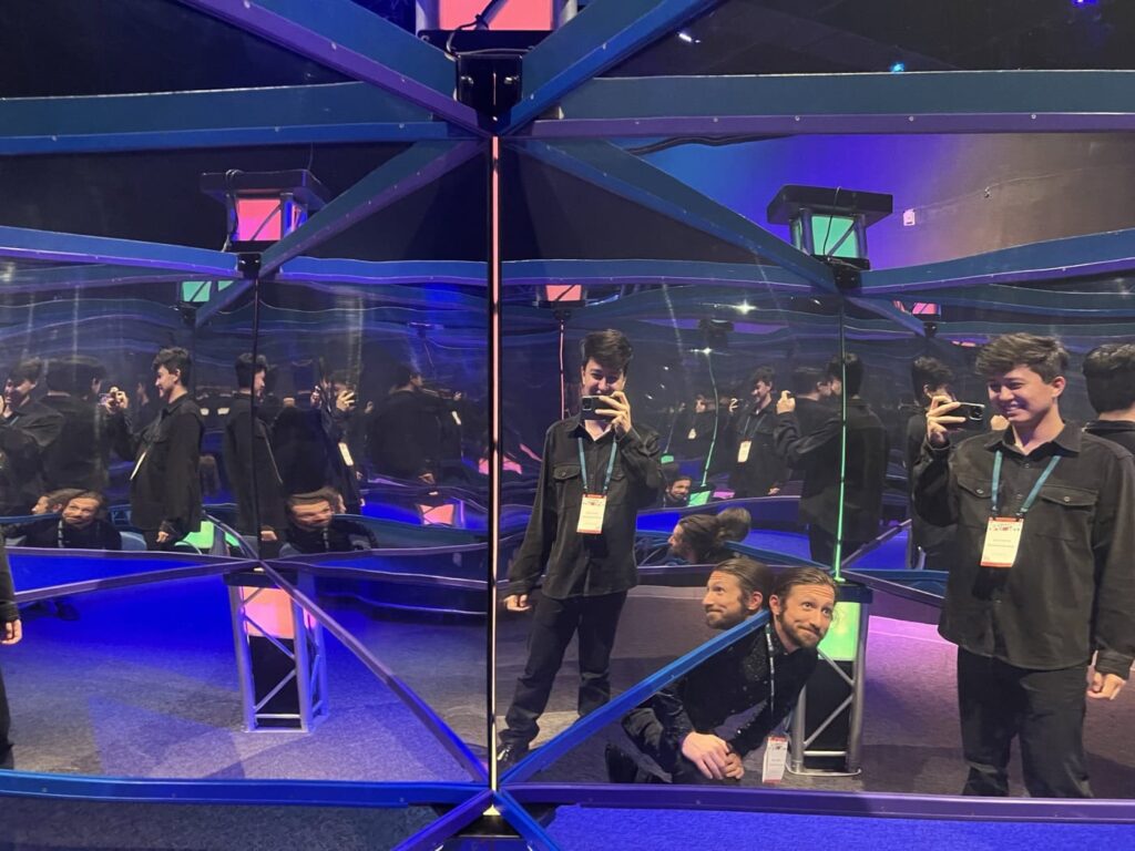 Vinícius and Max inside of a chamber filled with mirrors, creating multiple reflections in all directions. The space is illuminated with vibrant blue and pink lights.