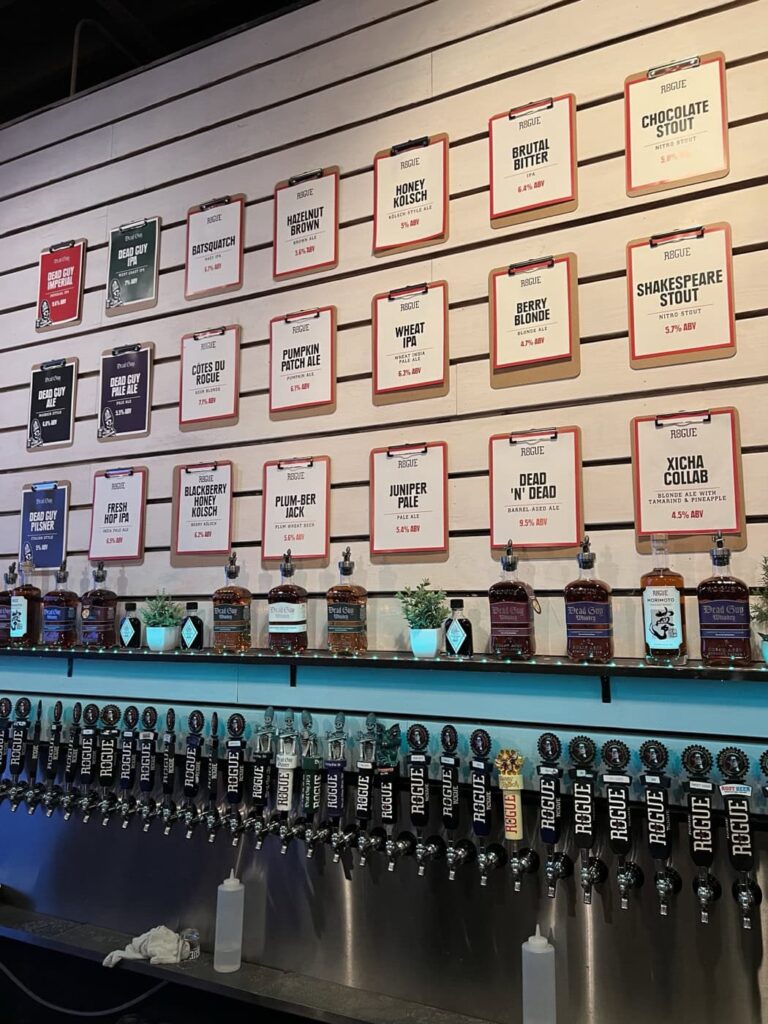 Rogue Brewery's wall showcasing different beer options on clipboards. Below the signs is a row of beer taps labeled with the brewery's logo, along with small plants and bottles of spirits decorating the counter.