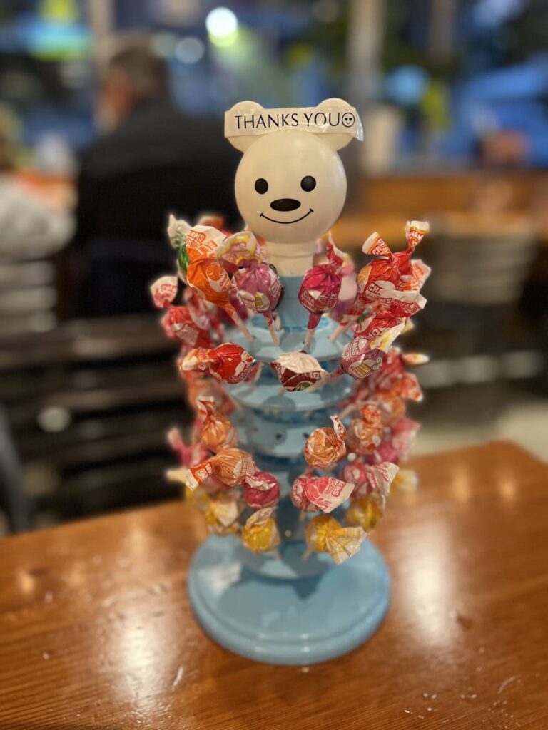 A light blue lollipop holder topped with a smiling bear head sits on a table at Wu-Rons ramen restaurant with a variety of colorfully wrapped suckers sticking out. A small label on top reads THANKS YOU.