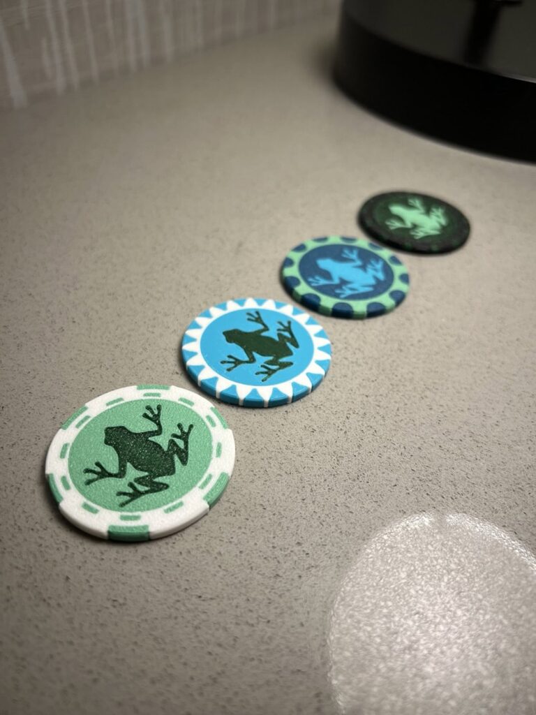 Four 3D printed poker chips are lined up on a gray surface. Each chip has a frog silhouette in the center. The chips are in various colors: green, blue, and black, with distinct patterns around the edges.