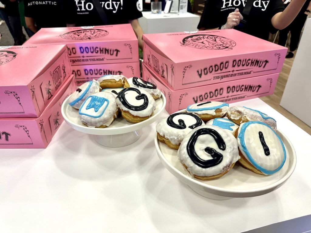 Plates of doughnuts with white icing and black and blue letters are displayed on a table. Behind them are stacks of pink boxes labeled "Voodoo Doughnut." People in black T-shirts stand in the background.