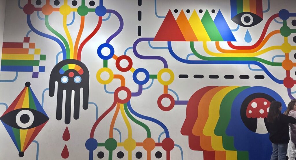 A vibrant mural with abstract shapes, bold lines, and rainbow colors. Features include eyes, overlapping rainbow-colored human profiles, and connected circles with flowing lines, creating a visually dynamic composition on the wall.