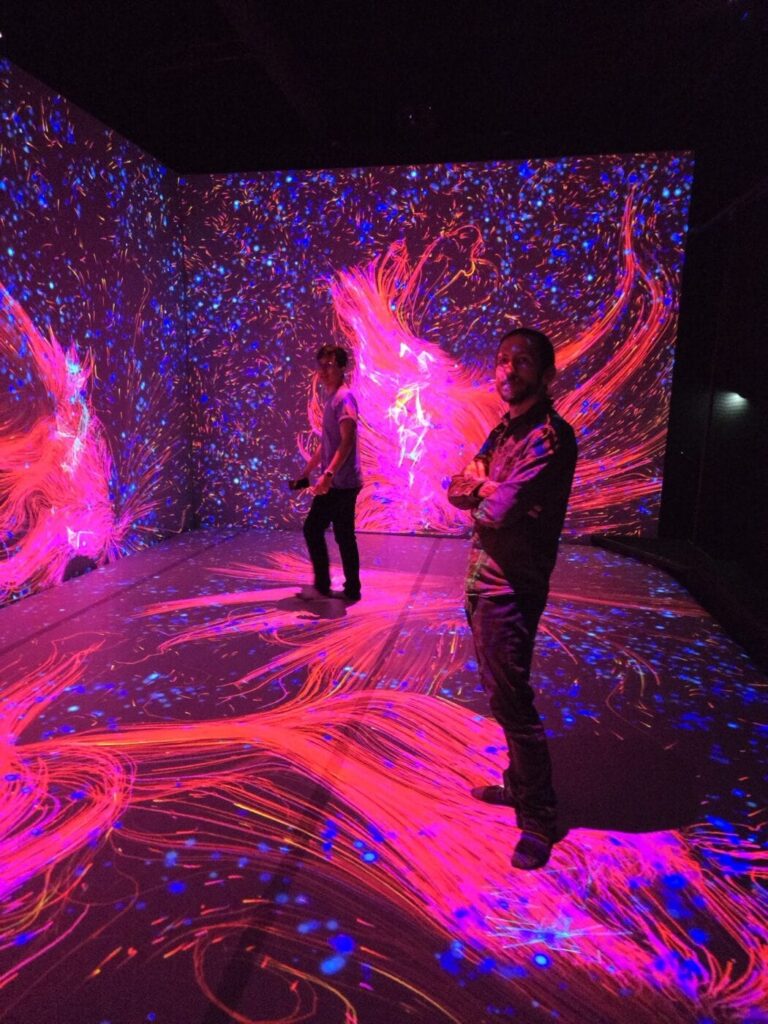 Max and Vinícius in a dark room illuminated by vibrant, swirling patterns of pink, blue, and orange light on the walls and floor, creating an immersive, digital art experience.